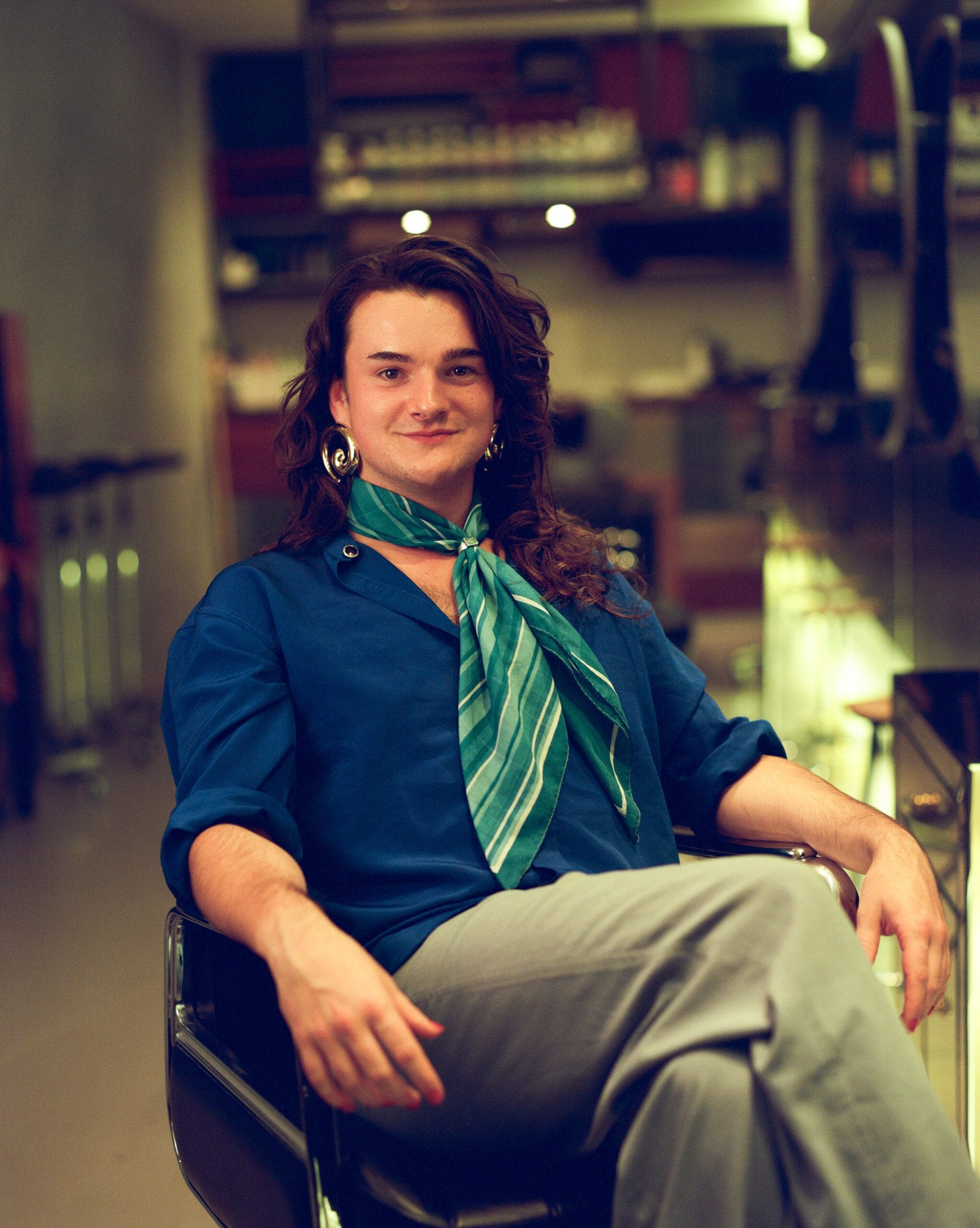 Persoon die in kappersstoel zich en je lachend aankijkt. Deze persoon heeft sjaaltje en zet met de benen gekruist over elkaar heen.