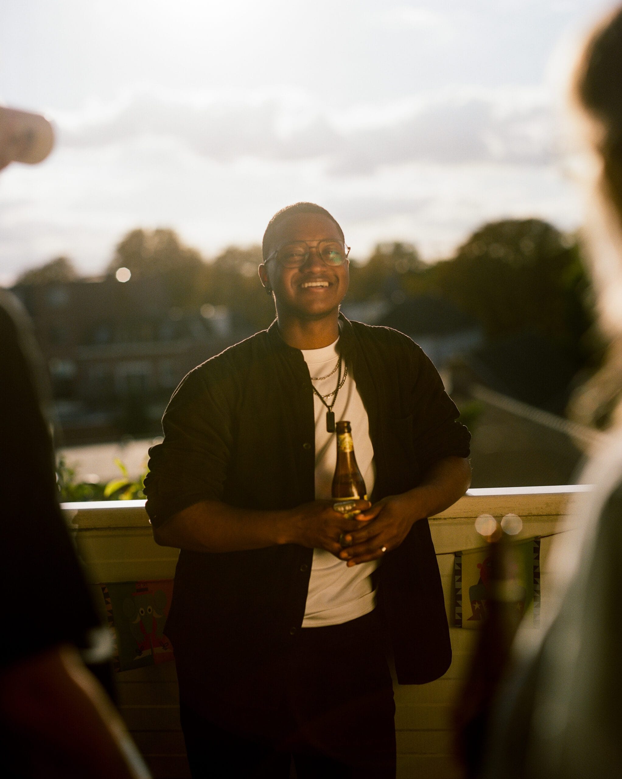 Je ziet een persoon op een balkon staan, met een biertje in de hand, die lacht naar de camera.